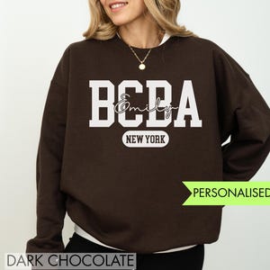 Op de afbeelding: Donkerbruine sweatshirt met witte blokletters "BCBA" boven de naam "Emily" in script. Daaronder staat "NEW YORK" in een witte ovaal. De woorden "PERSONALISED TEXT" staan in een groene banner. De woorden "DARK CHOCOLATE" staan onderaan.