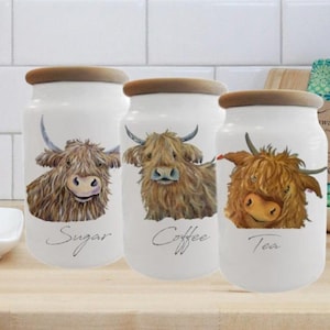 May include: Set of three white ceramic canisters with brown lids. Each canister features a different Highland Cow illustration and the words "Sugar", "Coffee", and "Tea" written in cursive.