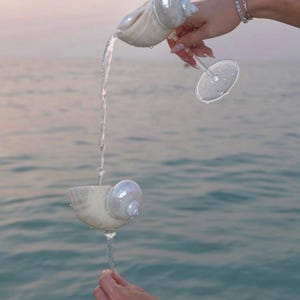 May include: A champagne glass and a seashell-shaped vessel are used to pour water. The glass is held by a person with a pearl bracelet. The water flows into the glass, creating a visually appealing display against the backdrop of the sea.