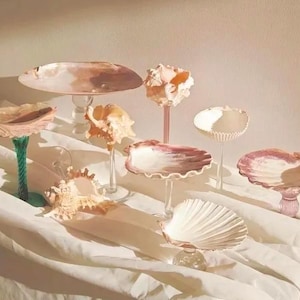May include: A collection of decorative seashell-shaped serving dishes and pedestals in various sizes and colours. The collection includes a large, round pedestal dish and smaller shell-shaped bowls with pink and white hues.