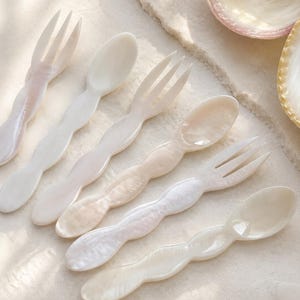 Pode incluir: Um conjunto de seis pequenos utensílios feitos de madrepérola branca iridescente, incluindo três garfos e três colheres. Cada peça tem um design de cabo ondulado e suave com acabamento brilhante, exibindo variações naturais de cor.