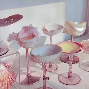 Könnte beinhalten: Eine Sammlung von rosafarbenen Weingläsern, jedes mit einer Schale aus einer natürlichen Muschel. Die Muscheln variieren in Form und Farbe, von blassrosa bis cremeweiß und gelb. Die Stiele und Basen sind in einem passenden Rosaton gehalten.