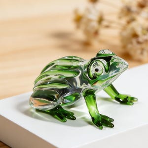 以下が含まれることがあります： 緑と透明な色合いの、詳細なガラス製のカエル置物。カエルは白い表面に置かれ、脚を伸ばしています。置物は光沢のある仕上がりで、背景はぼやけています。