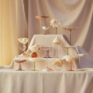 May include: A collection of decorative glass and seashell objects, including stemmed glasses and small dishes, arranged on a draped surface. The pieces feature a variety of colors, including pink, orange, and white, and are illuminated by soft lighting.