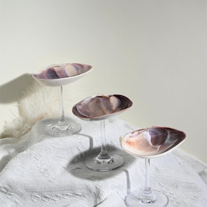 Könnte beinhalten: Drei dekorative Muschelschalen auf transparenten Glasstielen. Die Muscheln haben eine natürliche Farbpalette aus Weiß, Lila und Braun. Sie sind auf einer weißen, strukturierten Oberfläche angeordnet und bilden eine elegante Präsentation.