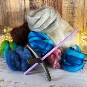 May include: A collection of colorful wool roving and fiber, including shades of blue, green, brown, and gray. A purple and green spindle is in the foreground. The materials are arranged on a white wooden surface, suggesting a craft or textile theme.