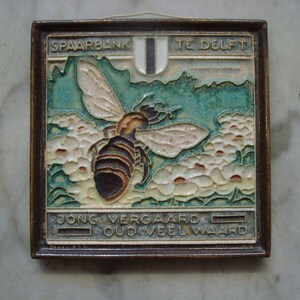 May include: A framed ceramic tile with a bee design. The bee is brown and black with cream wings, set against a backdrop of stylized flowers and a teal landscape. The text "SPAARBANK TE DELFT" and "JONG VERGAARD OUD VEEL WAARD" are visible.