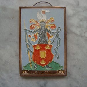 Op de afbeelding: Een keramische tegel met een wapenschild waarop een ridder met een speer en een schild in rood en geel te zien is. De tegel is omlijst met hout en heeft de tekst "KLEVE" onder het wapenschild.