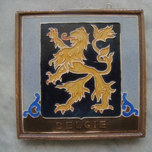 Könnte beinhalten: Eine braune Keramikfliese mit einem blauen und goldenen Löwenemblem und dem Wort "BELGIE" in goldenen Buchstaben.