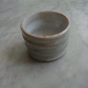 Może przedstawiać: Mały, biały kubek ceramiczny o lekko fakturowanej powierzchni. Kubek jest pusty i ma zaokrąglony kształt.