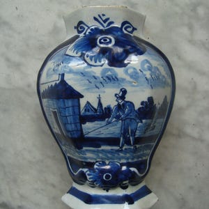 Könnte beinhalten: Eine blau-weiße Keramikvase mit einer Landschaftsszene. Die Vase zeigt eine detaillierte Malerei eines Mannes, der in der Nähe eines Gebäudes fischt. Die Vase hat eine einzigartige Form und dekorative florale Akzente.