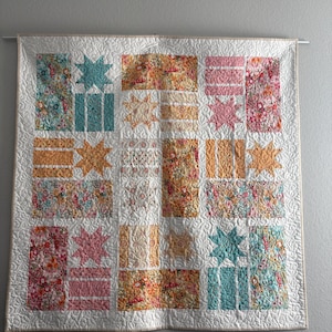 May include: A floral patchwork quilt with a white background, featuring squares and star shapes in shades of pink, orange, and teal. The quilt is hanging on a wall, showcasing its intricate design and craftsmanship.