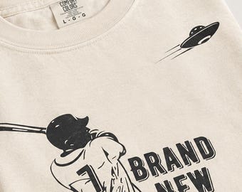 Może przedstawiać: Kremowy t-shirt z czarną grafiką przedstawiającą baseballistę zamachującego się kijem, z numerem 7 na koszulce, napisem "BRAND NEW" w postarzanym stylu oraz latającym spodkiem. Koszulka posiada metkę marki Comfort Colors.