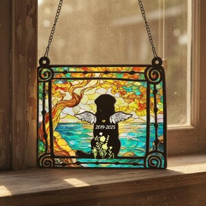 May include: Stained glass window art featuring a black silhouette of a dog with angel wings, the years 2019-2025, and floral accents. The background depicts a colorful landscape with a tree, water, and sky, all framed by a black border and hanging chain.