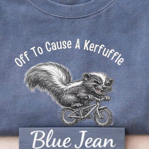 May include: Blue jean colored t-shirt with the text "Off To Cause A Kerfuffle" and a skunk riding a bicycle. The shirt has a crew neck and a small tag at the top. The words "Blue Jean" are on a small wooden sign.