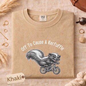 May include: Khaki-colored sweatshirt with the text "Off To Cause A Kerfuffle" and a graphic of a skunk riding a bicycle. The sweatshirt has a crew neck and a tag that says "Comfort Colors".