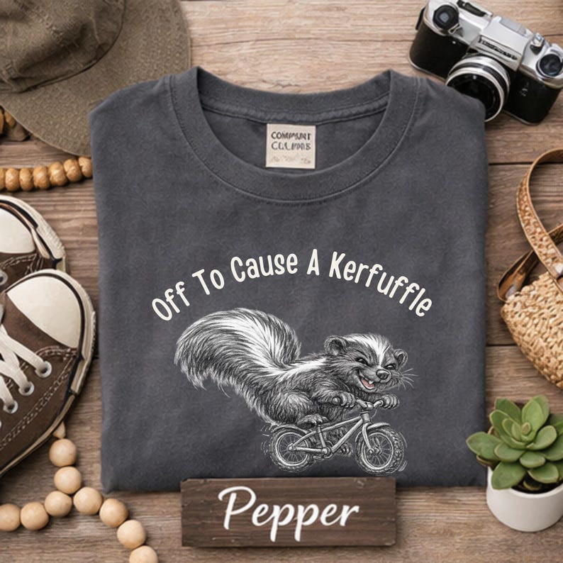 May include: Dark gray t-shirt with the text "Off To Cause A Kerfuffle" and a skunk riding a bicycle. The word "Pepper" is printed on a small wooden sign. A camera, hat, shoes, and a small plant are in the background.