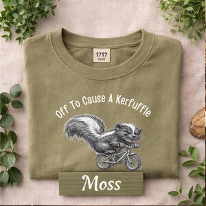 May include: Olive green t-shirt with a graphic of a skunk riding a bicycle. The text "Off To Cause A Kerfuffle" is above the skunk, and the word "Moss" is below. The shirt has a crew neck.