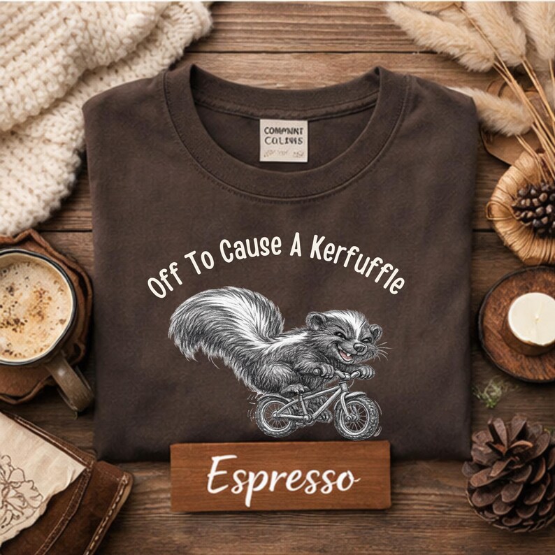 May include: Espresso-colored t-shirt with the phrase "Off To Cause A Kerfuffle" and a skunk riding a bicycle. The shirt has a crew neck and a tag on the upper back. A wooden sign with the word "Espresso" is at the bottom.