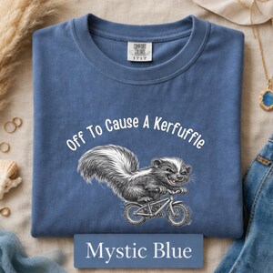 May include: Mystic blue t-shirt with the text "Off To Cause A Kerfuffle" above a skunk riding a bicycle. The shirt is a Comfort Colors 1717. The text "Mystic Blue" is on a rectangular label at the bottom.