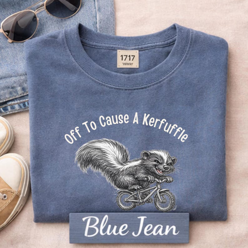 May include: Blue jean colored t-shirt with the text "Off To Cause A Kerfuffle" and a skunk riding a bicycle. The shirt has a crew neck and a small tag at the top. The words "Blue Jean" are on a small wooden sign.