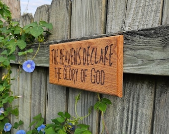 May include: A rectangular wooden sign with the text "THE HEAVENS DECLARE THE GLORY OF GOD" in a dark font. The sign is mounted on a weathered wooden fence, with green vines and blue flowers in the background.