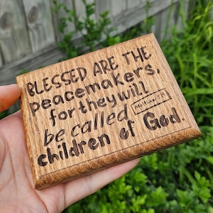 May include: A wooden plaque with the engraved text "BLESSED ARE THE peacemakers, for they will be called children of God." The text is in a dark brown color. The plaque also has the text "Matthew 5:9".