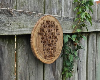 May include: A wooden plaque with a natural wood grain finish, featuring engraved text from Galatians. The plaque is oval-shaped and mounted on a weathered wooden fence. Green vines grow along the fence, adding a natural element.
