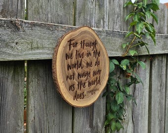 May include: A round wooden plaque with a natural bark edge, engraved with a quote from 2 Corinthians 10:3. The plaque is mounted on a weathered wooden fence, with green foliage in the background.