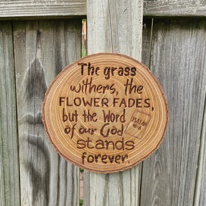 May include: A round wooden plaque with the text "The grass withers, the flower fades, but the Word of our God stands forever" and the reference "Isaiah 40:8" is burned into the wood. The plaque is displayed against a weathered wooden fence.
