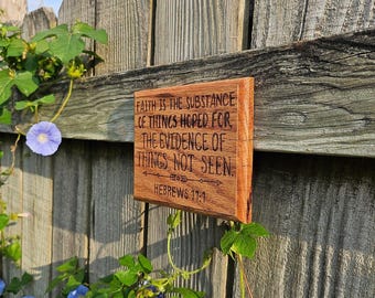May include: A wooden sign with the text "FAITH IS THE SUBSTANCE OF THINGS HOPED FOR THE EVIDENCE OF THINGS NOT SEEN" and "HEBREWS 11:1" engraved on it. The sign is rectangular and mounted on a wooden fence, with green foliage and blue flowers in the background.