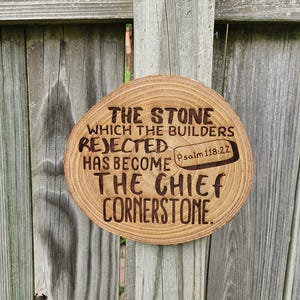 May include: A round wooden plaque with the text "THE STONE WHICH THE BUILDERS REJECTED HAS BECOME THE CHIEF CORNERSTONE" and "Psalm 118:22" etched into the wood. The plaque is a light brown color and is hanging on a wooden fence.