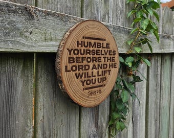 May include: A round wooden sign with the engraved text "Humble yourselves before the Lord and He will lift you up." The sign is made from a cross-section of a tree trunk and is mounted on a wooden fence.