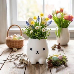 Könnte beinhalten: Ein weißer Keramik-Hasen-Blumentopf gefüllt mit gelben Narzissen, blauen Traubenhyazinthen und weißen Gänseblümchen. Ein Weidenkorb mit bunten Ostereiern und eine Vase mit Tulpen sind ebenfalls vorhanden.