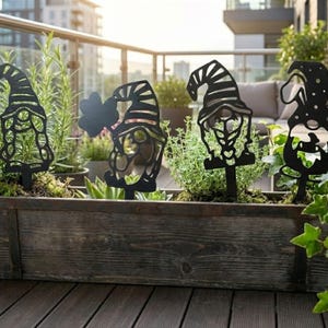 Könnte beinhalten: Schwarze Metall-Gartenzwerge in verschiedenen Designs, darunter ein Zwerg mit einem vierblättrigen Kleeblatt, werden in einem hölzernen Pflanzkasten mit Grünzeug ausgestellt. Die Zwerge sind auf Stäben, perfekt für die Gartendekoration.