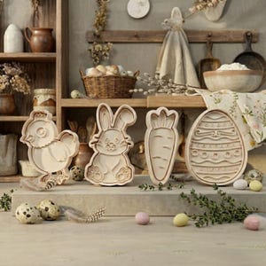 Puede incluir: Cuatro cortadores de galletas de Pascua de madera: un pollito, un conejito, una zanahoria y un huevo. Los cortadores de color marrón claro están sobre una superficie clara, con huevos decorativos y ramitas de vegetación cerca. Una exhibición rústica con temática primaveral.