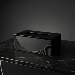 Puede incluir: Caja de pañuelos negra rectangular con un diseño curvo y texturizado en la parte delantera. La parte superior tiene una ranura estrecha para dispensar los pañuelos. La caja está hecha de un material oscuro y se encuentra sobre una superficie de mármol negro.