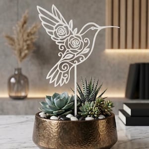 Puede incluir: Una estaca de planta de colibrí de metal blanco con detalles florales se coloca en una maceta texturizada de color bronce. La maceta contiene varias suculentas y pequeñas piedras. El fondo incluye un jarrón con plantas secas y un panel de madera.