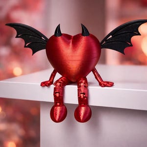 Peut inclure: Figurine rouge en forme de cœur avec des cornes et des ailes noires, des jambes et des pieds rouges, assise sur une surface blanche. La figurine a une apparence diabolique, avec une finition brillante. L'arrière-plan est flou avec un bokeh rose et blanc.