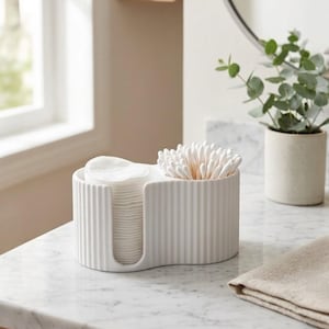 Può includere: Un organizer da bagno in ceramica bianca a coste che contiene dischetti di cotone e bastoncini. L'organizer è appoggiato su un piano in marmo, con una pianta e uno specchio sullo sfondo. Il design è moderno e minimalista.