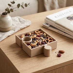 Può includere: Un vassoio per snack geometrico marrone chiaro pieno di una varietà di noci, pretzel e gocce di cioccolato. Il vassoio ha un piccolo scomparto circolare e un'estensione a forma di maniglia. Il vassoio è su una superficie di legno.