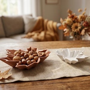 Può includere: Due ciotole a forma di foglia, una color terracotta e una bianca, sono posizionate su un runner testurizzato. La ciotola color terracotta è riempita con noci intere e fichi secchi. Sparse attorno alle ciotole ci sono foglie autunnali marroni e una ghianda.
