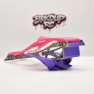 May include: A radio-controlled car chassis featuring a bright pink upper frame and a purple lower section with a metallic skeleton. The logo BRKDUP RC is visible in the background. The structure includes multiple small screws and metal plates.