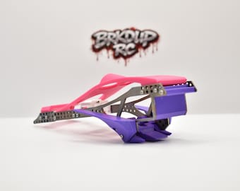 May include: A radio-controlled car chassis featuring a bright pink upper frame and a purple lower section with a metallic skeleton. The logo BRKDUP RC is visible in the background. The structure includes multiple small screws and metal plates.