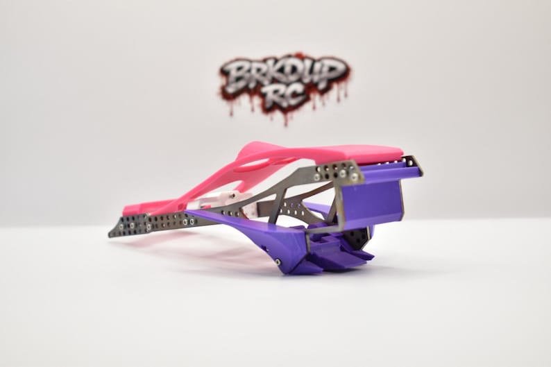 May include: A radio-controlled car chassis featuring a bright pink upper frame and a purple lower section with a metallic skeleton. The logo BRKDUP RC is visible in the background. The structure includes multiple small screws and metal plates.