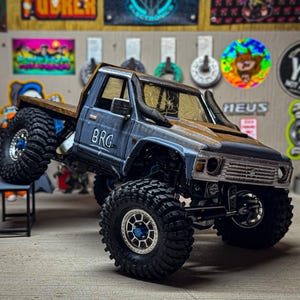 May include: A blue and brown RC truck with large, black off-road tires. The truck has the letters "BRG" on the side and is posed on a small black table. The background features various stickers and signs.
