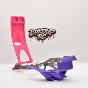 May include: A pink and purple RC car chassis stand featuring a tall vertical support with two circular cutouts and a horizontal base. The logo BRKDUP RC is displayed in a stylized font with a dripping effect above the stand.