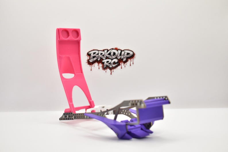 May include: A pink and purple RC car chassis stand featuring a tall vertical support with two circular cutouts and a horizontal base. The logo BRKDUP RC is displayed in a stylized font with a dripping effect above the stand.