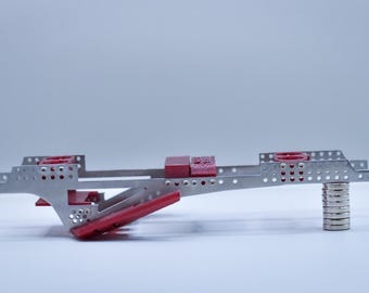 May include: A metallic construction frame featuring a series of perforated silver beams and plates. The structure includes several red plastic components attached to the frame, with a stack of circular silver magnets supporting one end of the assembly.
