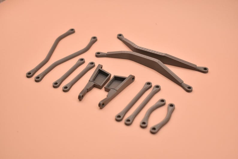 May include: A collection of dark gray, matte-finished plastic components for a remote-controlled vehicle, including two long curved suspension arms, two straight support bars, two angled chassis mounts, and four short curved links.